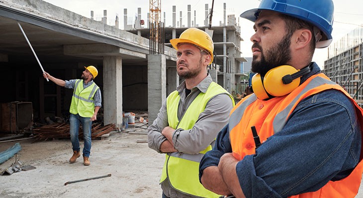 © pressmaster - Adobe Stock Zwei Kollegen mit Schutzhelmen und -westen stehen auf einer Baustelle, während im Hintergrund ein weiterer Mann steht.